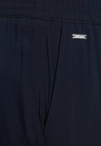 Mörkblått tyg med en slät textur och subtila veck. Inkluderar en liten metallisk logotag som är fäst vid sömmen.