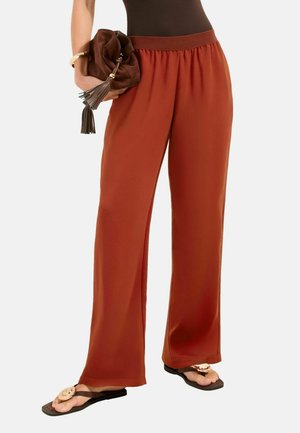 Femeie purtând pantaloni largi de culoare ruginie, ținând o geantă de mână maro cu franjuri și purtând sandale maro cu accente circulare în formă de floare.