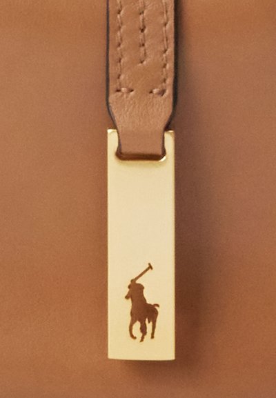 Gouden rechthoekige metalen tag met een reliëf van een polo-speler die een knuppel vasthoudt, bevestigd aan een gestikte lichtbruine leren band op een bruine leren ondergrond.