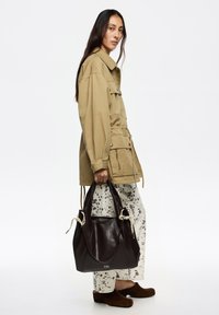 Brązowa skórzana torebka z węzłami z liny, trzymana przez osobę woversized beżowej kurtce i kremowych spodniach w kropki, w brązowych butach.