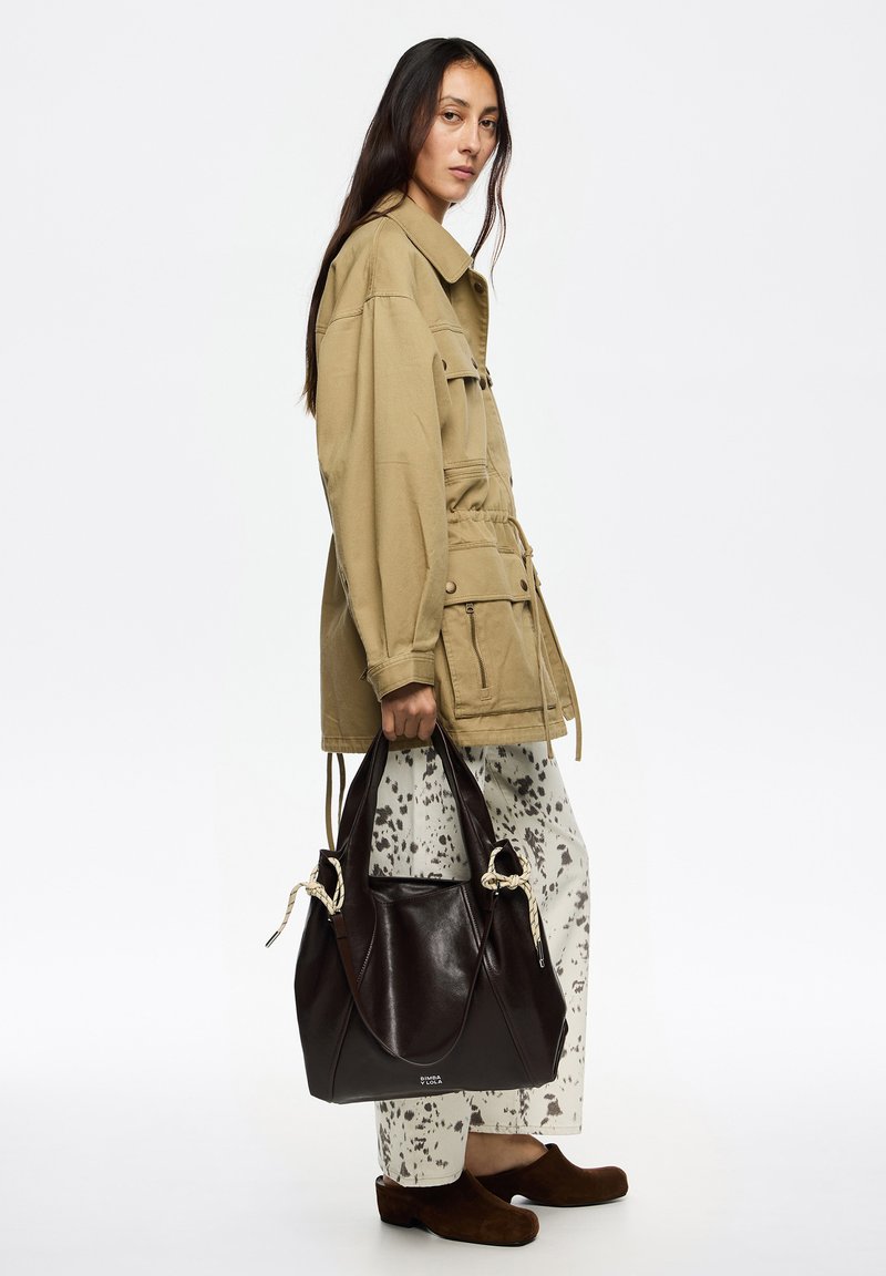 Brązowa skórzana torebka z węzłami z liny, trzymana przez osobę woversized beżowej kurtce i kremowych spodniach w kropki, w brązowych butach.