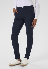 Navy slim-fit pants with front pockets, made of smooth fabric. Paired with white sneakers featuring a beige and black accent.
