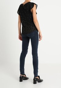Blusa negra con mangas cortas y onduladas, y patrón de puntos dorados, combinada con jeans ajustados azul oscuro y zapatos de tacón negros.