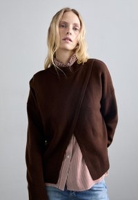 Rjav pleten pulover z visokim ovratnikom, ki ima čezpostavljen dizajn in rebraste robove, oblečen čez rdečo in belo črtasto srajco s ovratnikom.