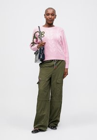 Różowy sweter z warkoczowym splotem i zielonymi haftami kwiatowymi, w zestawieniu z luźnymi oliwkowymi spodniami cargo z bocznymi kieszeniami. Czarna torebka i płaskie buty.