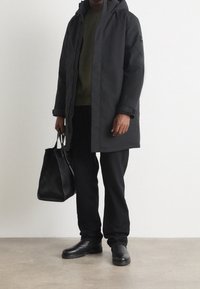 Svart isolerad parkas med huva, bärs över en mörkgrön tröja. Kombinerad med svarta byxor och stiliga svarta stövlar, håller en strukturerad svart väska.