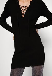 Svart ribbad klänning med en djup snörning i halsringningen. Långa ärmar och en strukturerad yta kompletterar dess smala, förlängda form.