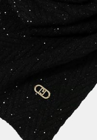 Tessuto nero a maglia con piccoli paillettes sparse e una spilletta ovale in metallo dorato con logo attaccata vicino al centro.