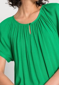 Grøn bluse med rund halsudskæring, bløde folder og en lille nøglehul-detalje, lavet af let tekstureret stof.