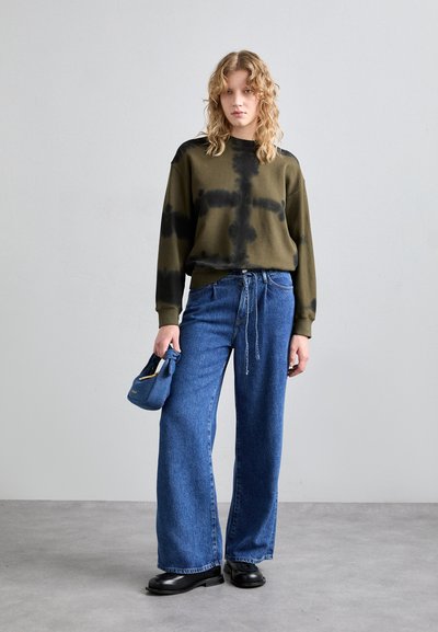 Hanorac tie-dye de culoare verde măslin, blugi largi din denim albastru cu șnur, pantofi negri și o geantă mică albastră.