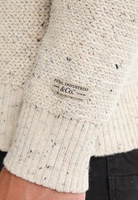 Maglione color crema realizzato in tessuto lavorato a maglia con polsini a coste. Presenta piccole macchie nere e blu in tutto il capo. L'etichetta recita "PE10L INDUSTRIES."