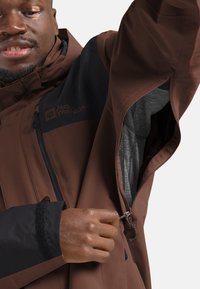 Braune wasserdichte Jacke mit schwarzen Akzenten, Reißverschlusstaschen und strukturiertem Innenfutter. Verfügt über verstellbare Manschetten und ein Logo auf der Brust.
