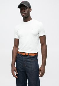 Λευκό κοντομάνικο μπλουζάκι με μικρό λογότυπο στο στήθος, συνδυασμένο με σκούρο μπλε τζιν και καφέ δερμάτινη ζώνη. Το γκρι καπέλο ολοκληρώνει την εμφάνιση.