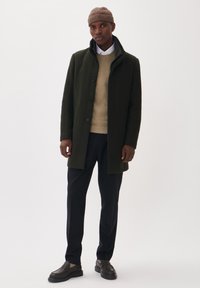 Manteau vert foncé avec un col haut et des boutons, porté sur un pull beige, associé à un pantalon noir et des chaussures sombres. Fond simple.