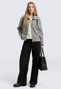 Grijs patroon zip-up jack met een puntige kraag, gecombineerd met een wit shirt en zwarte wijde broek. Inclusief zwarte handtas.