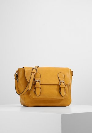 Żółta torebka na ramię w kolorze musztardowym z dwoma paskami z klamrami i regulowanym paskiem na ramię na białym piedestale na tle szarego tła.