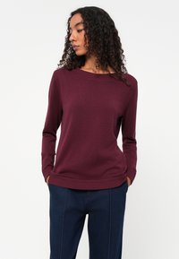 Pull en maille bordeaux avec un col rond et des poignets côtelés, assorti à un pantalon bleu marine, mettant en valeur une coupe décontractée et une texture douce.