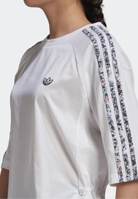 Vit atletisk tröja med rund halsringning, broderad logotyp och mönstrade tre-randiga accentlinjer på ärmarna. Sidodragkedja i nederkant.