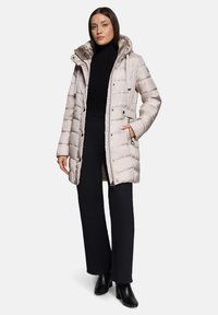 Manteau d'hiver matelassé beige avec capuche, présentant un motif matelassé, des poches avant et une fermeture éclair. Porté par-dessus un col roulé noir et un pantalon.