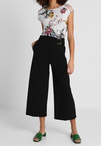 Biała bluzka w kwiatowy wzór z krótkimi rękawami, w połączeniu z czarnymi spodniami z szerokimi nogawkami i wysokim stanem. Modelka nosi zielone klapki.