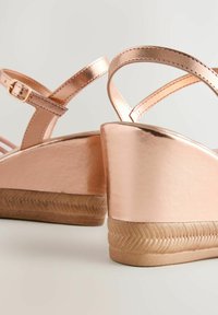 Sandalias cuña de color oro rosa con un acabado metálico, que presentan una base texturizada y una correa ajustable en el tobillo. Incluye acentos lisos y pulidos.