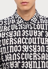 Black shirt featuring a repeating white text pattern with bold letters. Collar and buttoned front with black buttons. Smooth fabric texture.
