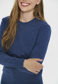 Žena se usmívá, má na sobě modrý svetr s malými metalickými hroty na hrudi, jemně zkřížené ruce přes trup.