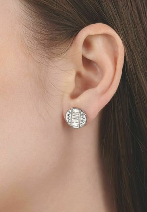 Oor van een vrouw met een ronde zilveren stekerearring met rechthoekige en kleine ronde heldere stenen in een symmetrisch patroon.