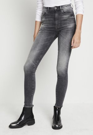 Personne portant un jean skinny noir délavé taille haute et des bottines noires, debout devant un fond clair uni.