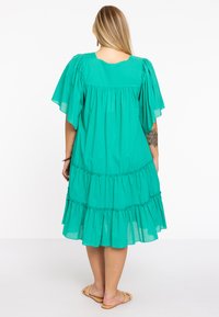 Groene katoenen jurk met korte spitse mouwen, gelaagde ruche aan de onderkant en een losse pasvorm. Achteraan is er een vierkante halslijn en geraffineerde stof te zien.