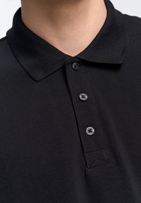 Juoda polo marškinėliai su tekstūruotu audiniu, trijų mygtukų apykaklė ir klasikinis krūtinės apykaklė. Yra logotipas mygtuko detalė ir siuvinėjimo akcentai.