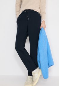 Personne portant un pull beige, un pantalon noir à cordon, des baskets blanches, tenant une veste polaire bleu clair sur un fond blanc.