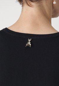 Top nero a coste con un charm a forma di insetto in metallo sulla parte posteriore del collo, che evidenzia l'artigianato e i dettagli di design unici.