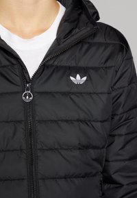 Svart puffjacka med horisontell sömnad, dragkedja fram och en liten vit logotyp på vänster bröst. Slätt, lätt material.