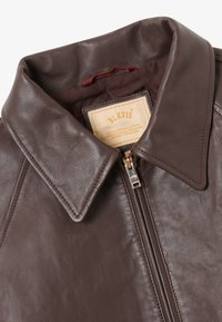 Hnědá kožená bunda s zaobleným límcem, zapínáním na zip a prošívanou podšívkou. Na štítku je nápis "BLKVIS" a design zahrnuje jemnou texturu.