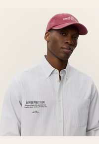 Chemise rayée bleue et blanche avec une poche poitrine, comportant le texte "LOWER WEST SIDE" et une casquette rose avec le même texte brodé.