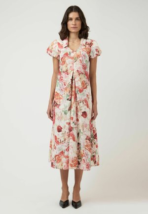 Kvinde med skulderlangt mørkt hår iført en knælængde blomsterkjole med pufærmer og sorte spidse sko, stående mod en ensfarvet baggrund.