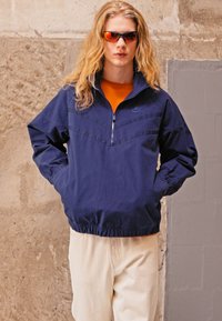 Tumši zils savilkšanas džemperis ar ceturtdaļas rāvējslēdzēju, elastīgām piedurknēm un jostasvietu. Uzmests virs oranžas krekla, kombinēts ar gaismas toņu biksēm.