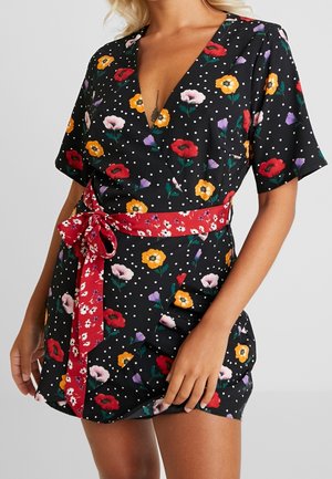 Kobieta ubrana w czarną, kwiecistą sukienkę kopertową w białe kropki z czerwonym kwiecistym paskiem w talii, prezentująca tatuaż na klatce piersiowej.