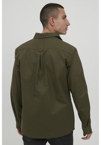 Camisa verde oscuro hecha de una tela resistente, con un pliegue en la espalda, puños con botón y un cuello puntiagudo. El diseño es de corte relajado.