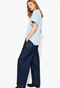 T-shirt oversized azzurro chiaro abbinato a pantaloni in denim larghi di un blu profondo. I pantaloni presentano cuciture minime e due tasche posteriori. Calzature nere.