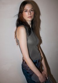 Szary top bez rękawów z delikatnym połyskiem, zestawiony z niebieskimi dżinsami. Modelka trzyma przezroczystą torbę. Tło ma jasny, neutralny odcień.