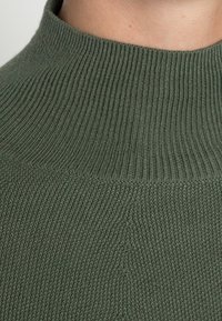 Grüner Strickpullover mit hohem geripptem Kragen und strukturiertem Gewebe, das eine nahtlose Verarbeitung und einen weichen, dehnbaren Stoff hervorhebt.