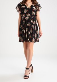 Robe noire à fleurs avec un décolleté en V, des manches courtes et volantes, et une jupe évasée. Présente des motifs floraux colorés et est associée à des sandales à talons noires.