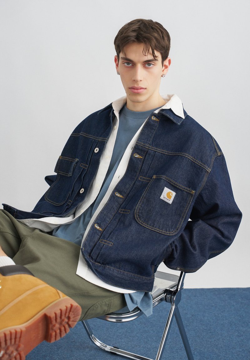 Junger Mann sitzt auf einem Metallstuhl und trägt eine blaue Jeansjacke mit weißem Kragen, ein blaues Hemd, olivgrüne Hosen und braune Stiefel.