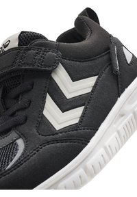Schwarze Sportschuhe mit strukturiertem Obermaterial, ausgestattet mit grauen Chevron-Akzenten, schwarzem Mesh und einer weißen Gummisohle. Klettverschluss für einfaches An- und Ausziehen.