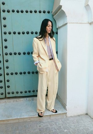 Mujer con traje color crema y sandalias negras está de pie con las manos en los bolsillos frente a una puerta color azul verdoso con tachuelas negras y pared blanca.