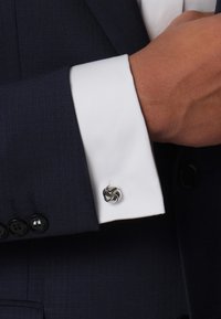Navy suit jacket cuff with a white shirt. Cuff features a twisted, silver cufflink and a smooth texture. Black buttons on the jacket are visible.