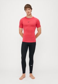 Rdeča kratka športna majica s kratkimi rokavi z majhnim črnim logotipom, v kombinaciji z oprijetimi črnimi legicami, ki na spodnjem delu hlačnic nosijo bel logotip.