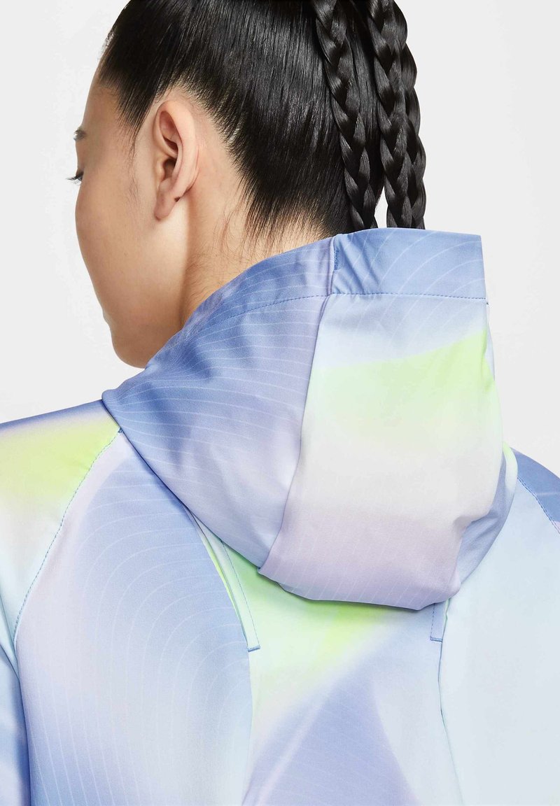 Hooded jack in een lichte stof met een verloop van pastelkleuren, vooral blauw met groene accenten, een gladde textuur en een subtiel gestreept patroon.